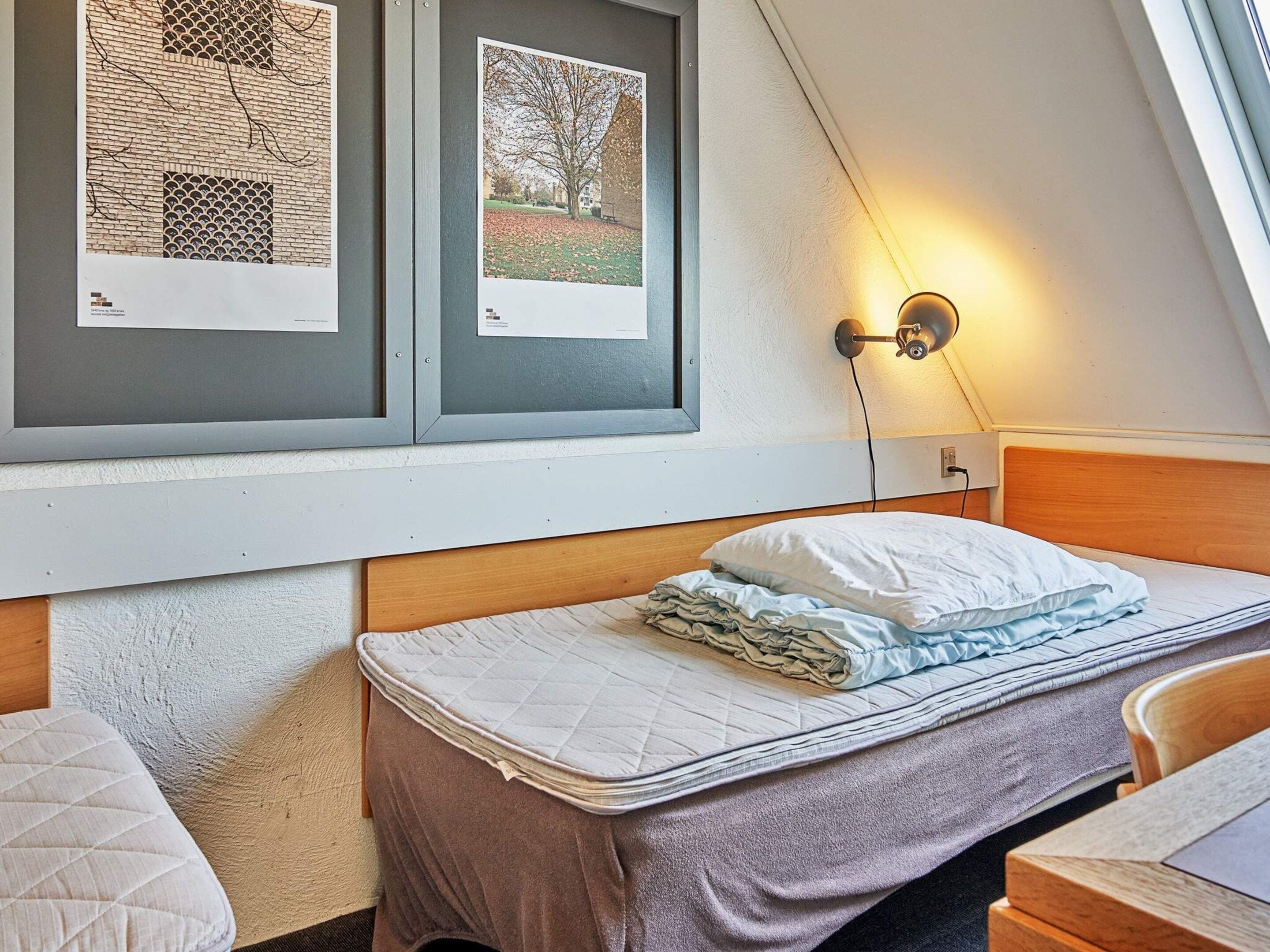 A bedroom with a bed, a nightstand, a wall-mounted lamp, and framed pictures on the wall.