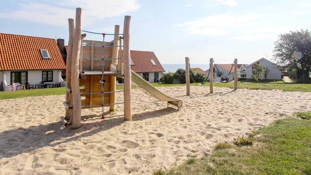 A playground with a wooden slide and climbing structure, set on sandy ground with houses in the background. - 5 Person Holiday Park Home in Gudhjem (Gudhjem)