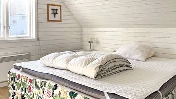 A bedroom with a sloped ceiling, a bed with a quilted mattress, a bedside table with a lamp, and a window with a view of the outside.