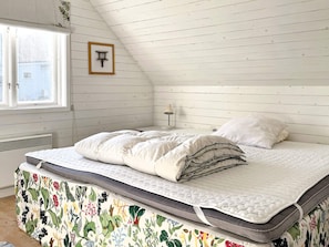 Un dormitorio con techo inclinado, cama con colchón acolchado, mesita de noche con lámpara y ventana con vistas al exterior.