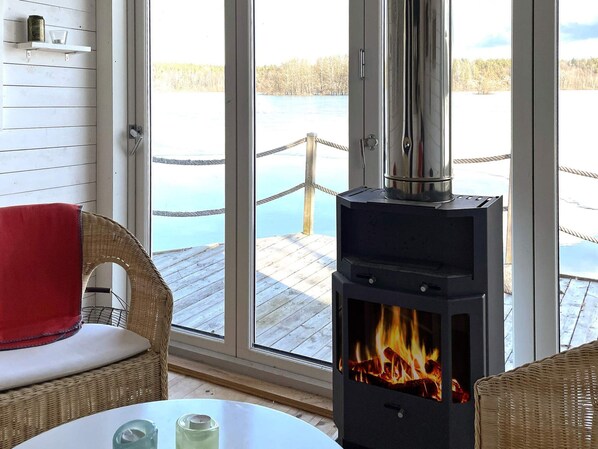 A room with a large window, a wicker chair, a table with candles, and a wood-burning stove.
