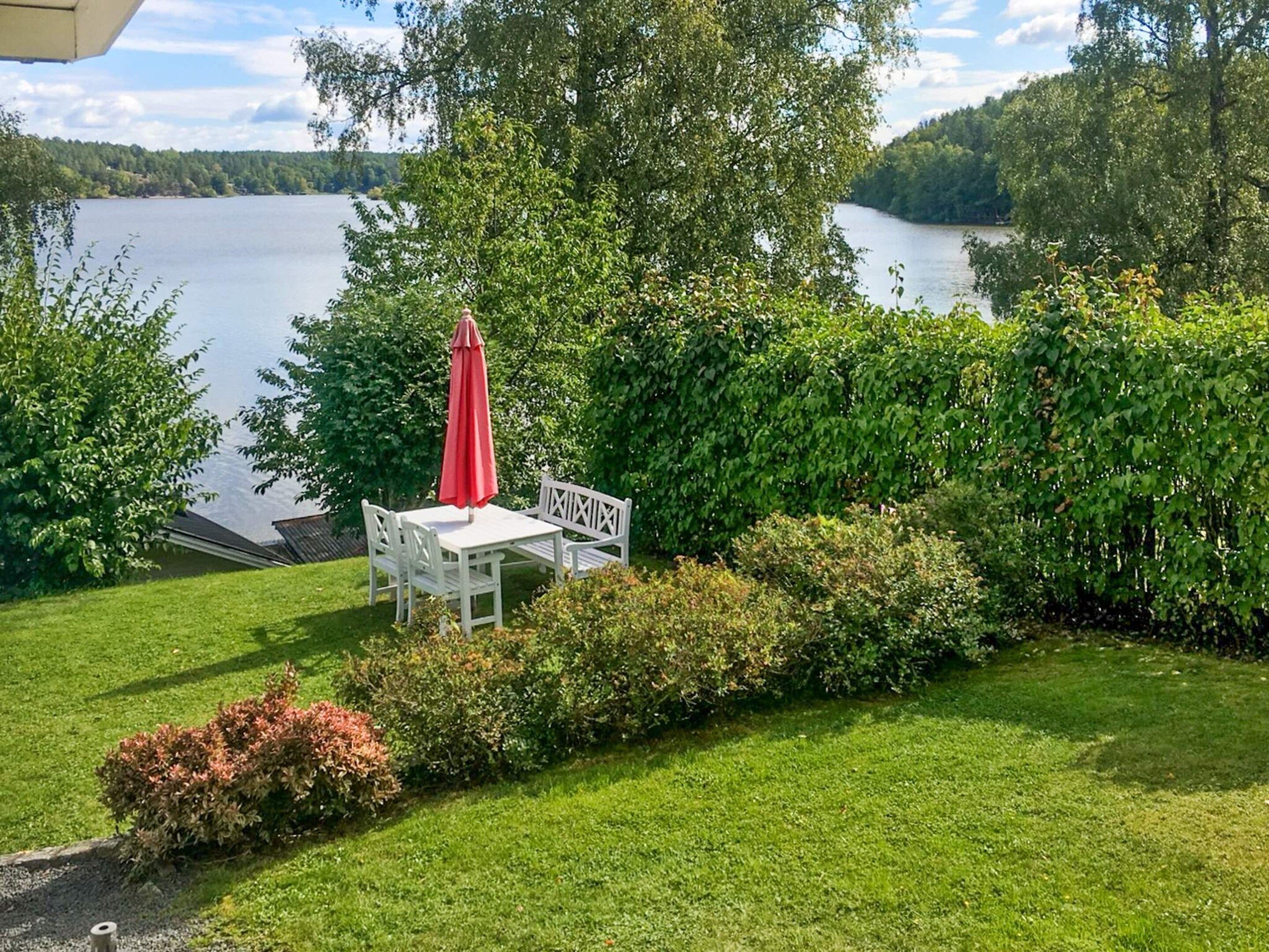 A patio with a table and chairs, a red umbrella, and a view of a lake.