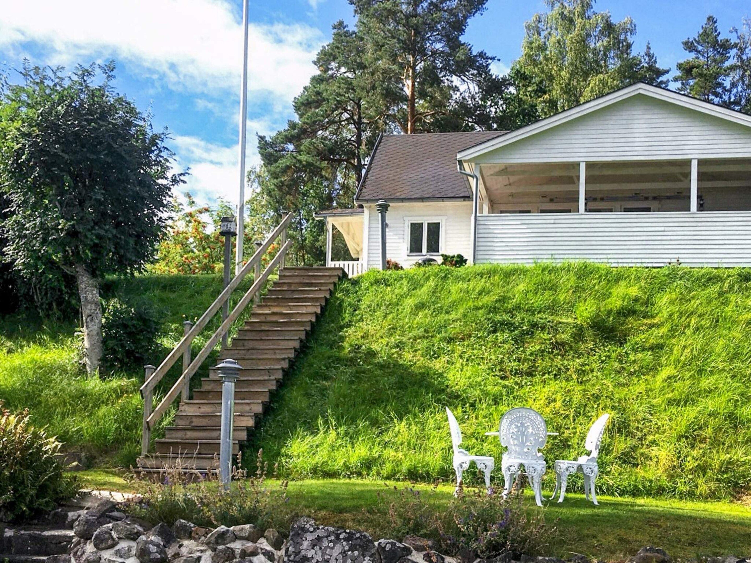 A house with a staircase leading to a covered porch, surrounded by greenery and a stone wall.