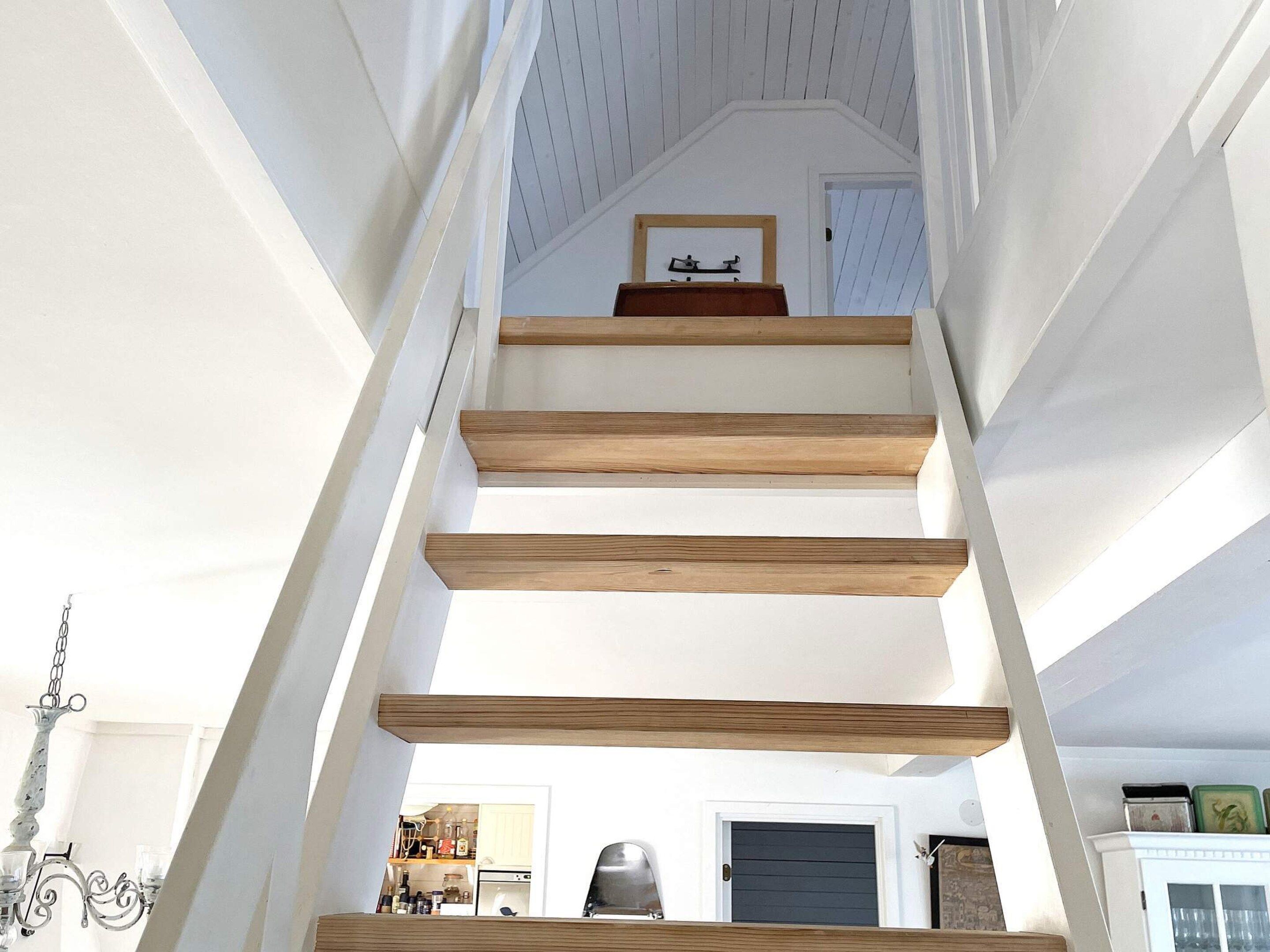 Una escalera con peldaños de madera y paredes blancas, que conduce a un rellano con un cuadro enmarco.