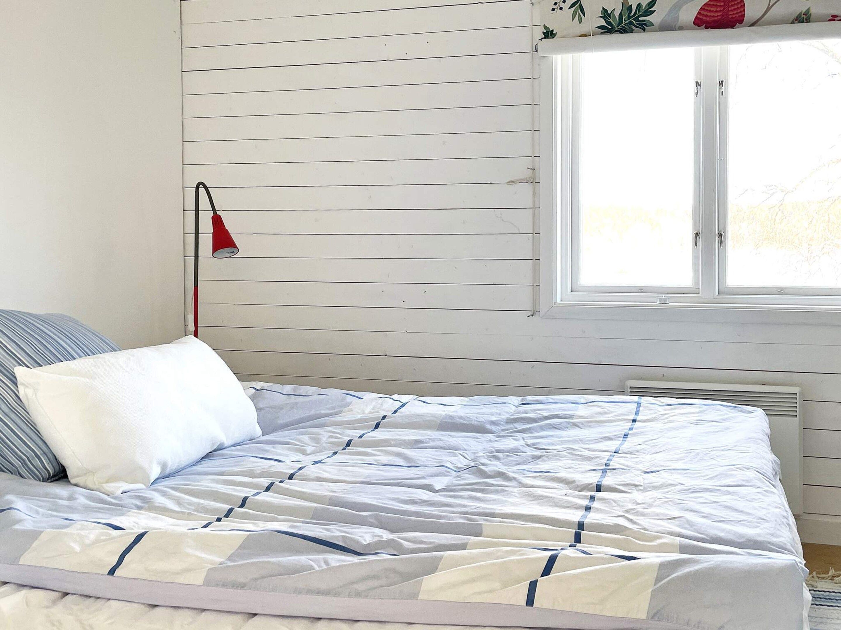 A bed with a striped pillow, a red lamp, and a window with blinds.