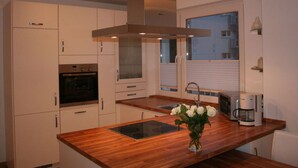 A modern kitchen with white cabinets, a built-in oven, a stainless steel range hood, a wooden countertop, a coffee maker, and a window with blinds. - Holiday Apartment Seega (Timmendorfer Strand)