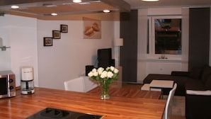A modern kitchen with a wooden countertop, a built-in stove, and a vase with white flowers. There is a dining area with a table and chairs, and a dark brown sofa in the background. - Holiday Apartment Seega (Timmendorfer Strand)