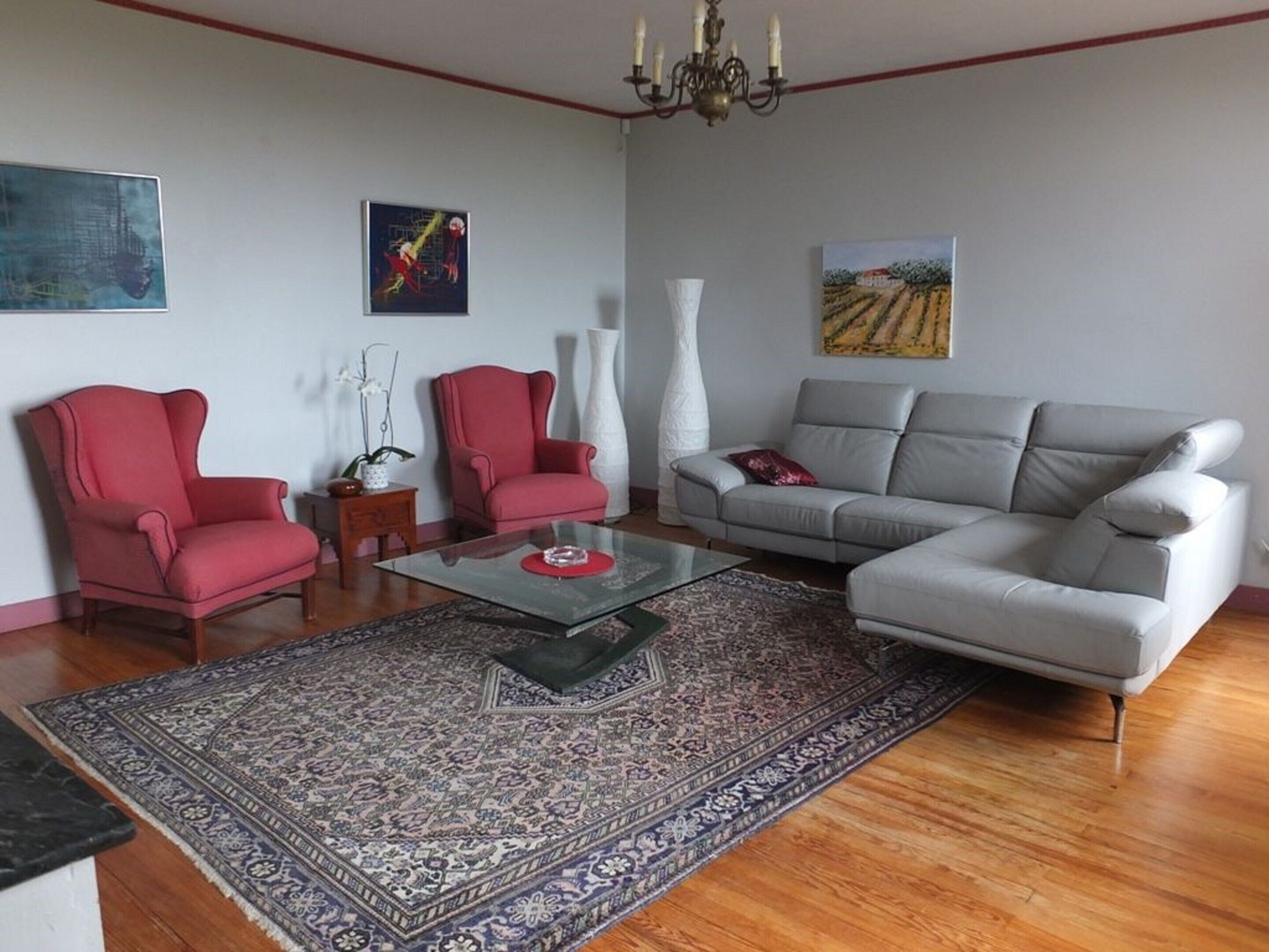 A living room with a grey sofa, two red armchairs, a glass coffee table, and a patterned rug.