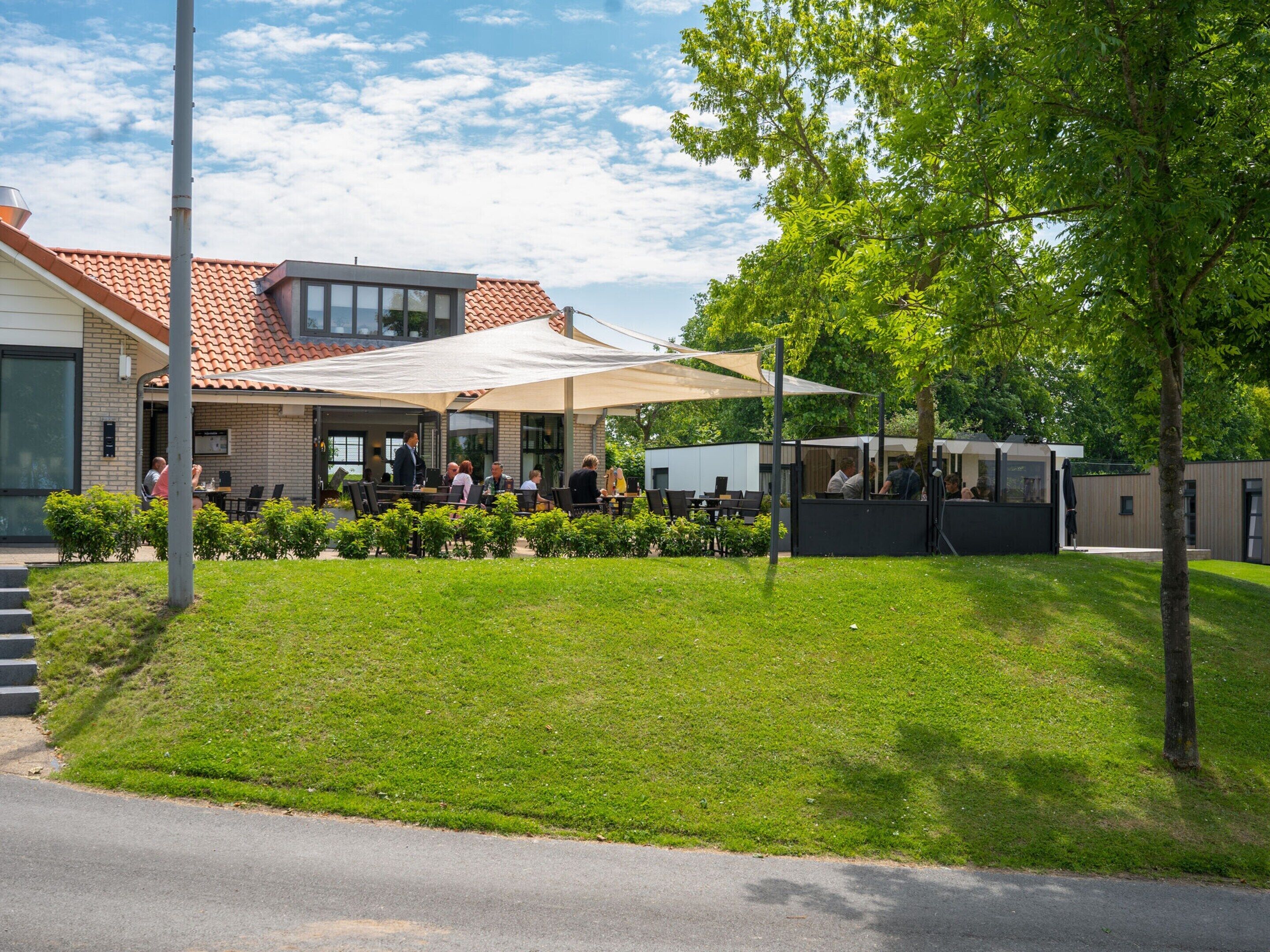 An outdoor cafe with tables and chairs, a building with a tiled roof, and a grassy area with trees.