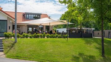 An outdoor cafe with tables and chairs, a building with a tiled roof, and a grassy area with trees.
