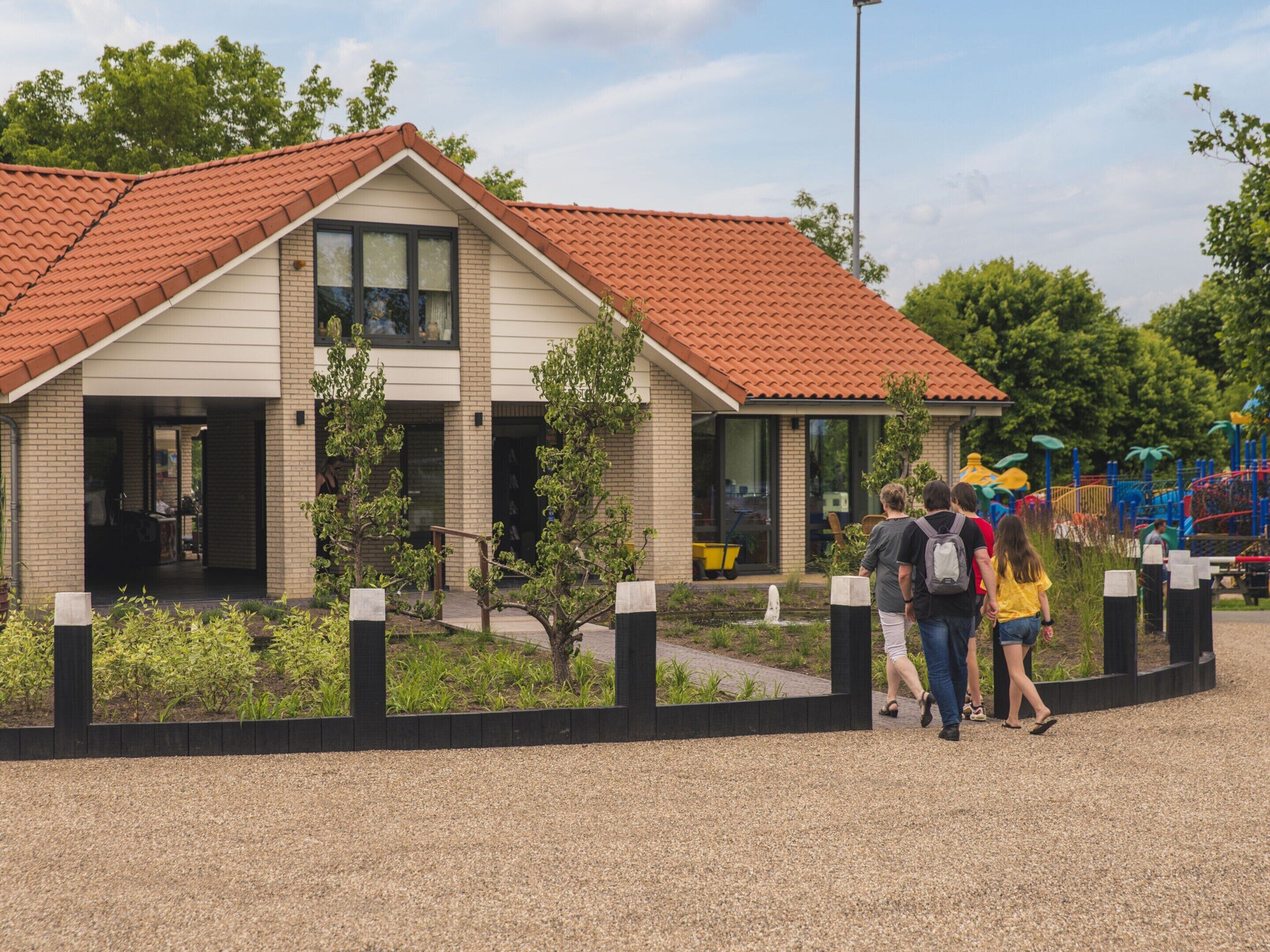 Een gebouw met een rode dakbedekking en een bakstenen gevel, omgeven door een grindpad en begroeiing.