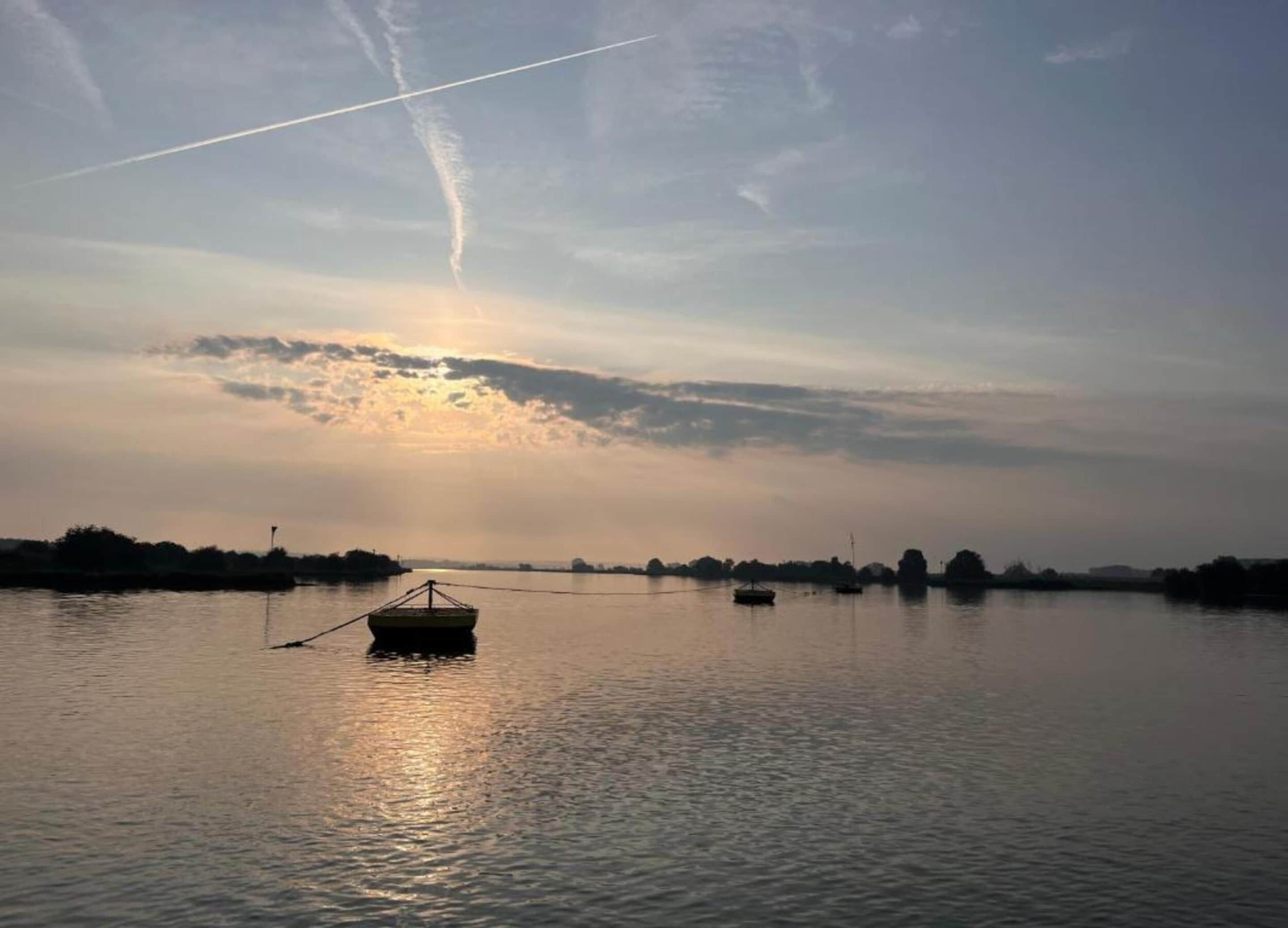 A calm body of water with a small boat anchored in the center, surrounded by a calm sky with a visible contrail.
