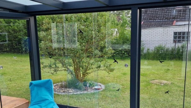 A modern room with a glass wall, a blue bean bag chair, and a view of a garden with a tree and birds. - Beach Chair No.1 Comfortable Vacation Home (Wangerland)