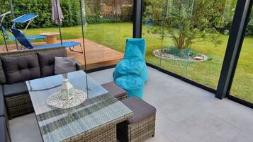 A modern outdoor living space with a glass table, wicker furniture, and a view of a garden.