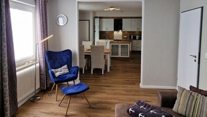 A modern living room with a blue armchair, a wooden floor, a dining area, and a kitchen in the background. - Beach Chair No.1 Comfortable Vacation Home (Wangerland)