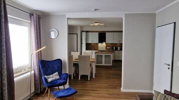 A modern living room with a blue armchair, a wooden floor, a dining area, and a kitchen in the background.
