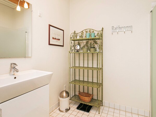 Casa de banho com lavatório branco, um quadro na parede, uma estante de metal com plantas em vasos e um contentor de lixo.