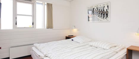 A bedroom with a bed, a window with curtains, a radiator, a wall-mounted picture, and a bedside table with a lamp.