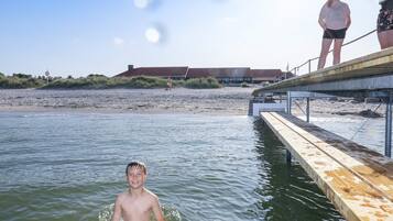 Un muelle de madera que se adentra en una masa de agua, con una playa arenosa y un edificio al fondo.