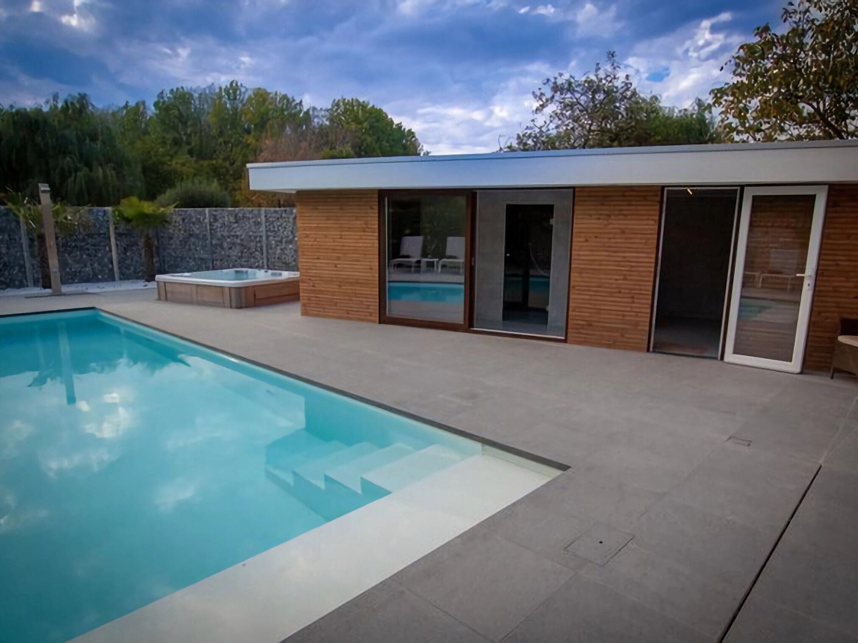 Un espace piscine extérieur moderne, doté d’une piscine à l’eau cristalline, d’un jacuzzi et d’un bâtiment en bois avec des portes vitrées.