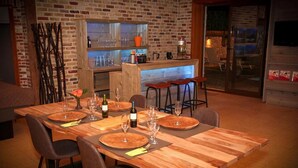 A dining area with a wooden table set for four, surrounded by chairs. A kitchen with a brick wall and bar stools is visible in the background. - Villa With Swimming Pool in Erembodegem (Aalst)