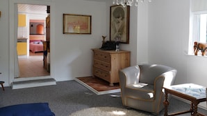 A room with a chandelier, a wooden dresser, a grey armchair, and a small table. There is a doorway leading to another room with a bed and a pink wall. - Holiday Home With art (Leipzig)