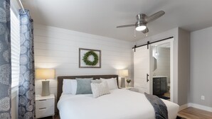Un dormitorio con cama, mesitas de noche, una lámpara de pared, ventilador de techo, una puerta y un espejo.