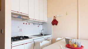 Eine kleine Küche mit weißen Schränken, einem Gasherd und einem Spülbecken. Ein Esstisch mit einer roten Untertasse und Gläsern ist ebenfalls vorhanden.