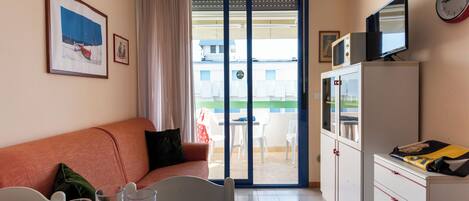 A room with a sofa, a dining table with glasses and a book, a cabinet with a TV, and a sliding glass door to the outside.