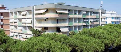 A multi-story building with balconies, surrounded by dense green trees.