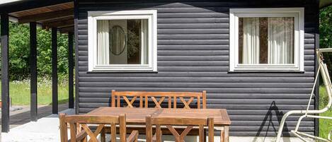 A cabin with two windows, a covered patio, and a table with chairs.