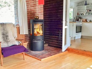 A cozy room with a wood-burning stove, a chair with a fur throw, and a kitchen area visible through an open door.