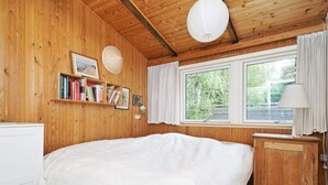A room with a bed, wooden walls, a window with curtains, a bedside lamp, and a shelf with books. - 6 Person Holiday Home in Rorvig (Rørvig)