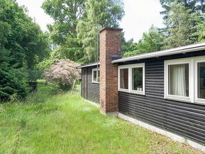Una casa con una chimenea de ladrillo, una ventana con cortina y un jardín con césped y árboles.