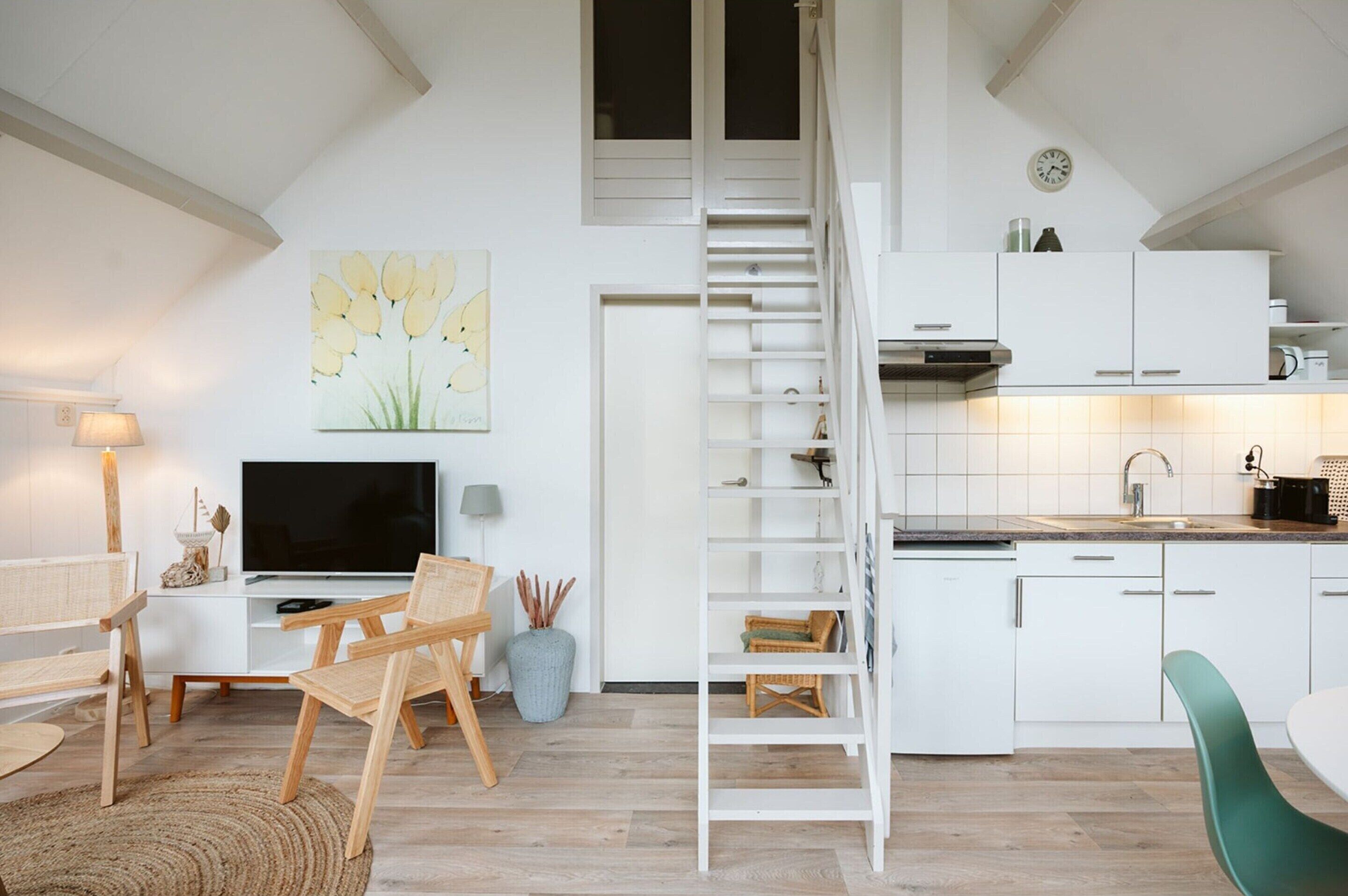 A modern kitchen with white cabinetry, a wooden ladder, a dining area with a green chair, and a TV on a white console table.