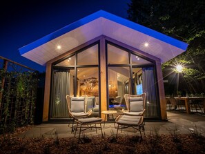 A modern, glass-enclosed structure with two wicker chairs and a small table, surrounded by a garden with mulch and plants. - Luxury Lodge With Private Swimming Pool (Rhenen)