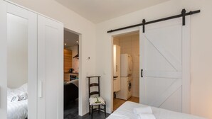 Ein Schlafzimmer mit Bett, Stuhl und Kleiderschrank. Durch eine offene Tür ist eine Küche zu sehen.