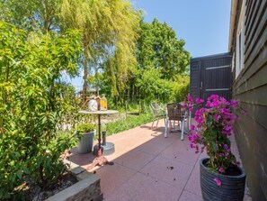 Une terrasse avec une table et des chaises, entourée de verdure et agrémentée d’un pot de fleurs aux fleurs roses.