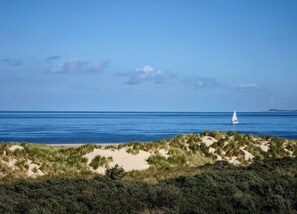 Een kustlandschap met een zandstrand, begroeide duinen en een zeilboot op het water.