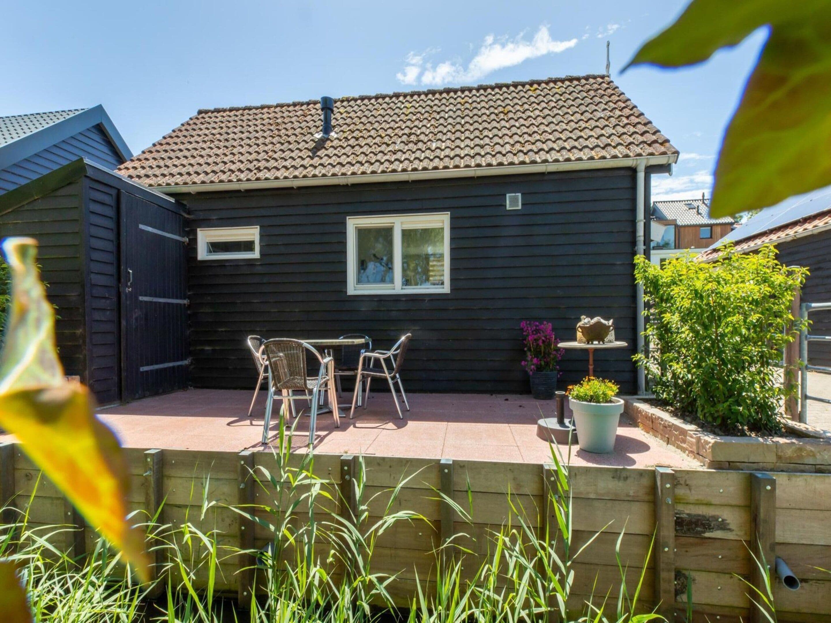 A residential property with a dark-colored exterior, a tiled roof, a fenced patio area with a table and chairs, and a small garden with plants and a decorative item.