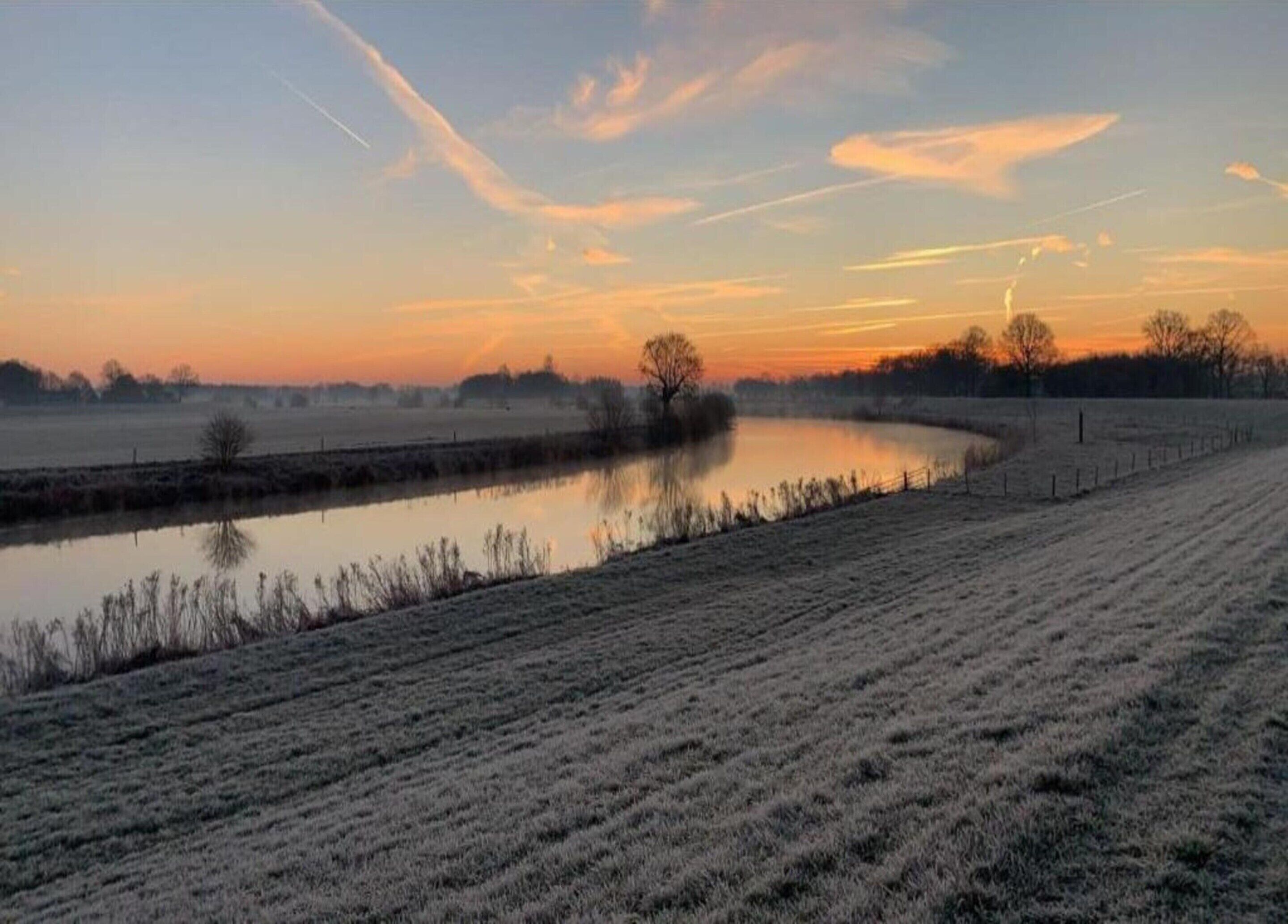 A frost-covered field with a calm water body and a fence in the foreground.