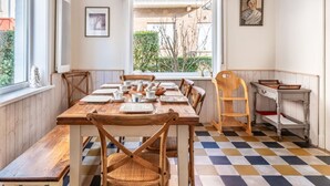 A dining area with a wooden table set for six, surrounded by chairs, a window with a view of greenery, and a small kitchen area with a cart.