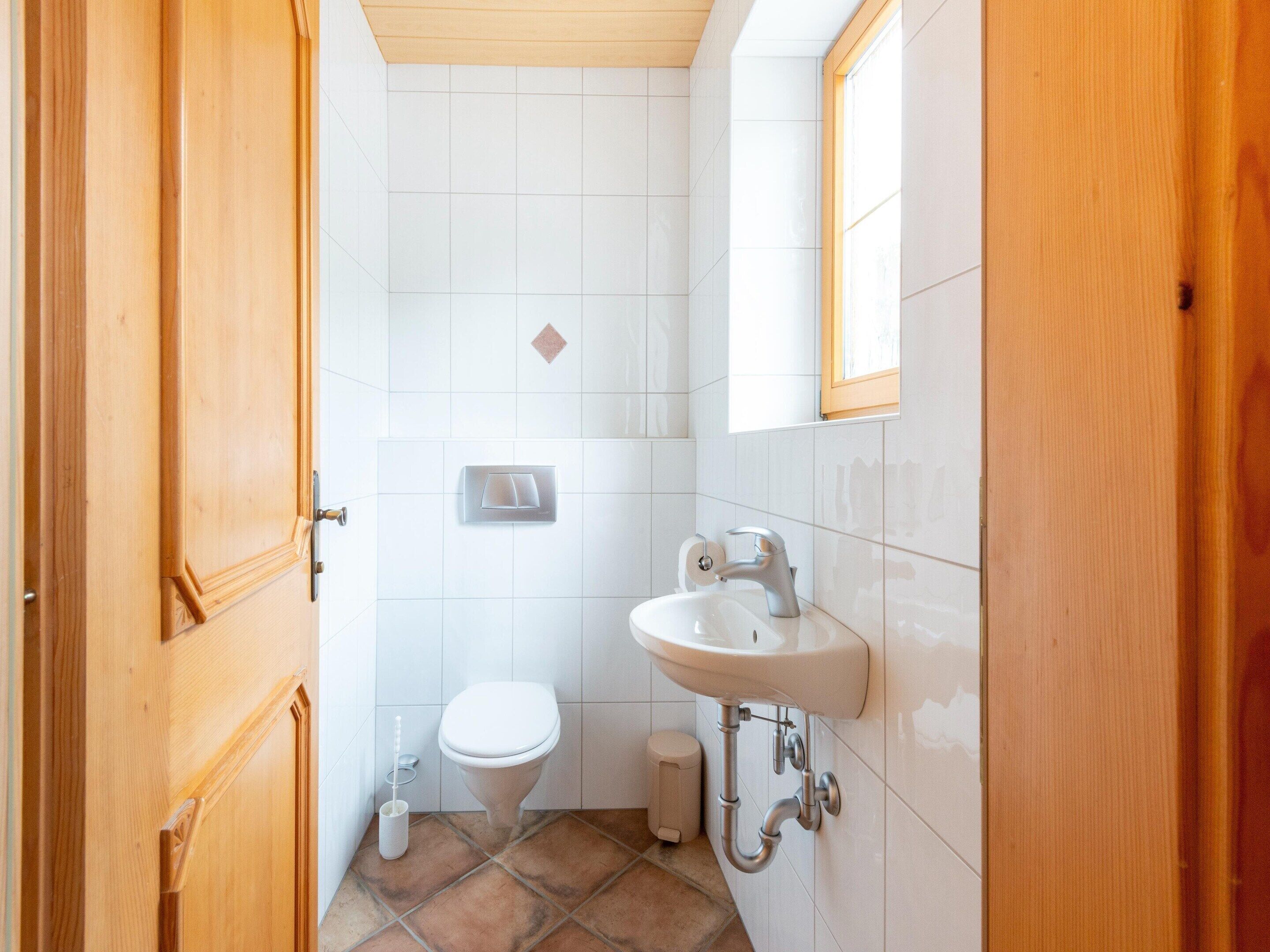 A small, clean bathroom with white tiles, a toilet, a sink, and a wooden door.