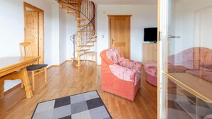 Uma sala de estar com escada espiral de madeira, uma mesa de jantar com cadeiras, um sofá e uma televisão sobre um móvel.