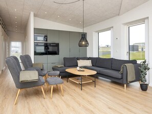 Un salon moderne avec un canapé gris, deux fauteuils, une table basse et un coin cuisine équipé d’un four à micro-ondes et d’un four traditionnel.