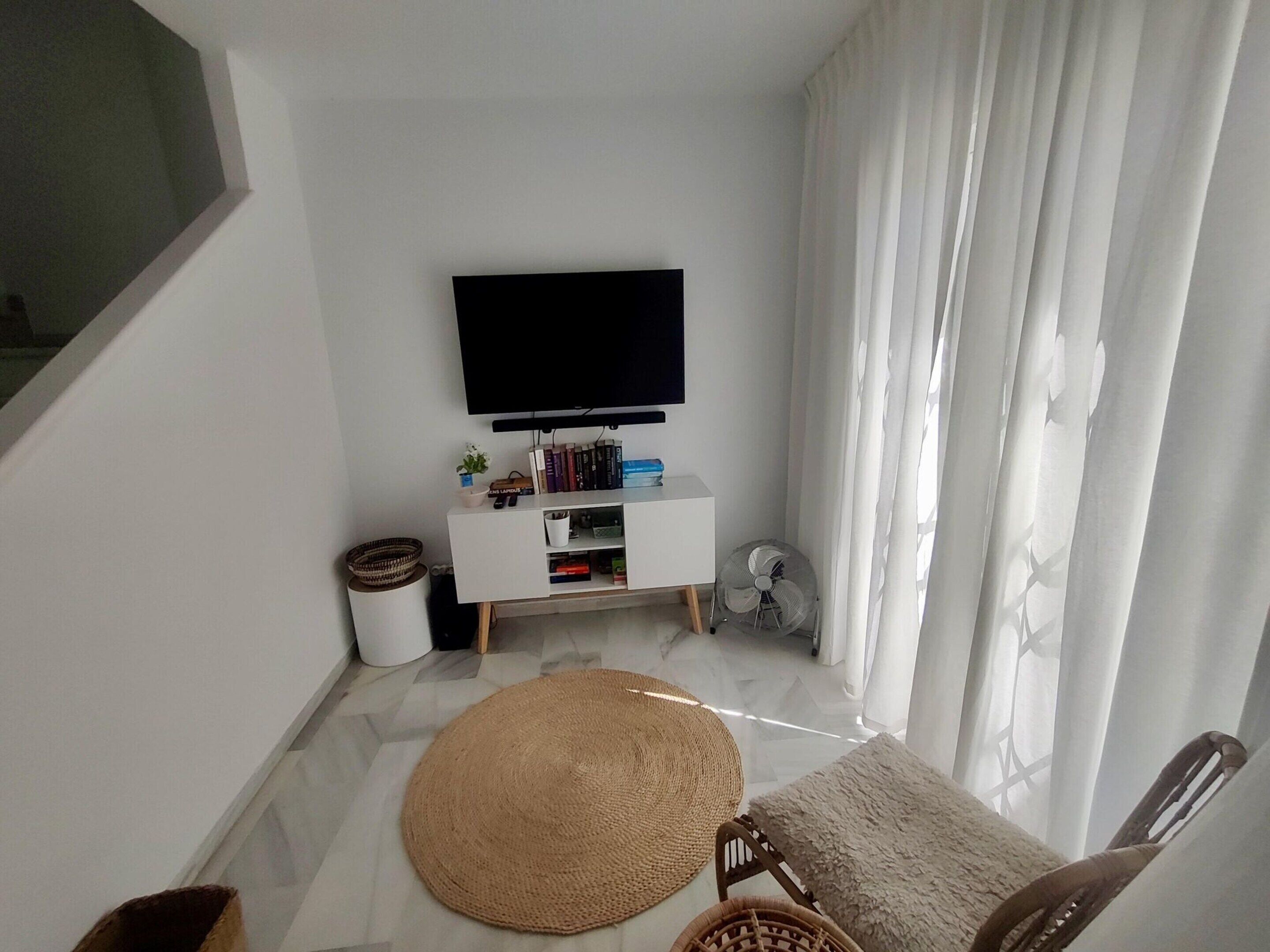 A living room with a flat-screen TV mounted on the wall, a white entertainment unit with books and decorative items, a woven rug, a wooden chair, and a white sofa.