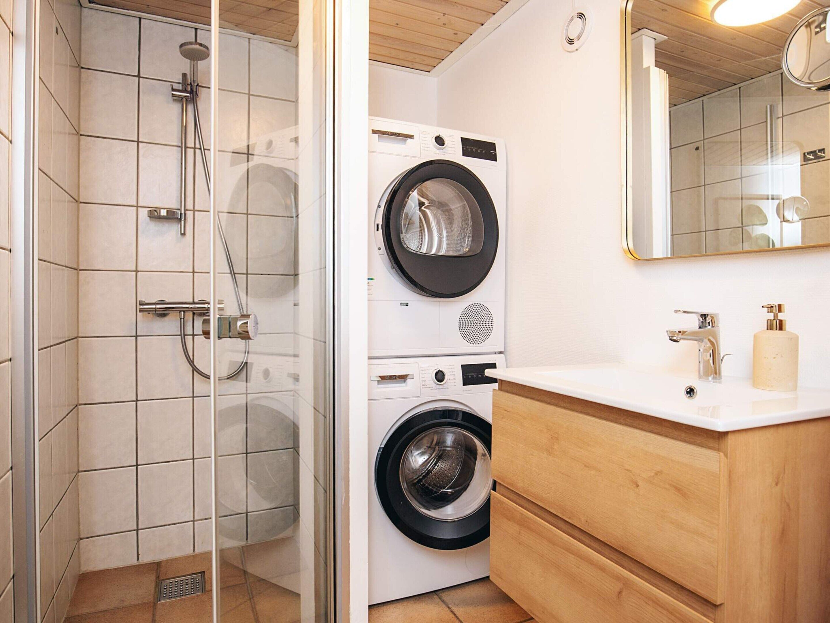 Une salle de bain avec une douche à parois de verre, un lave-linge et un sèche-linge superposés, un meuble-lavabo en bois et un miroir.