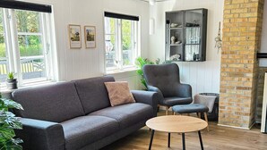 A modern living room with a grey sofa, a round wooden coffee table, and a grey armchair. There are two windows with curtains, a brick accent wall, and a black cabinet with decorative items.