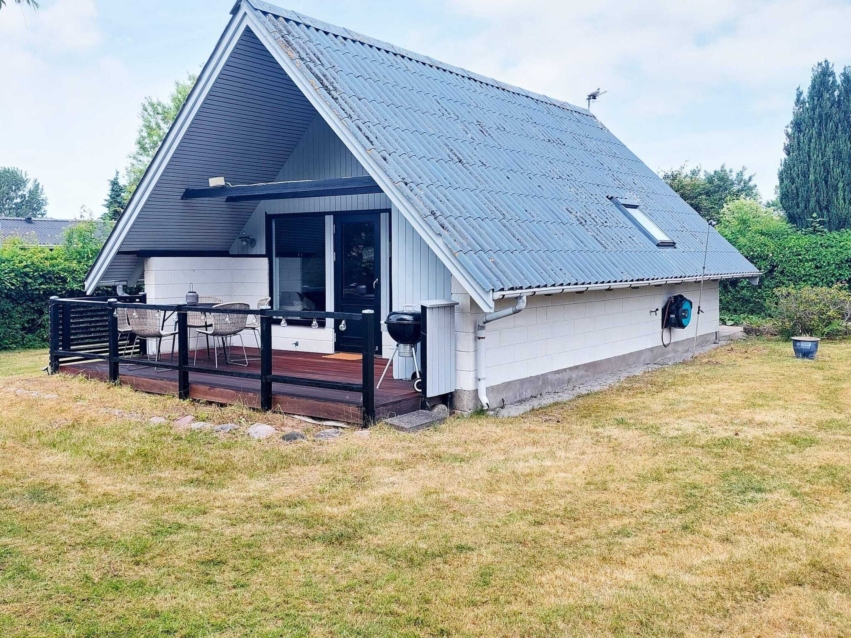 En enplansbyggnad med ett sluttande tak, en täckt veranda med utomhusmöbler och ett litet garage som är sammanbyggt med huset.