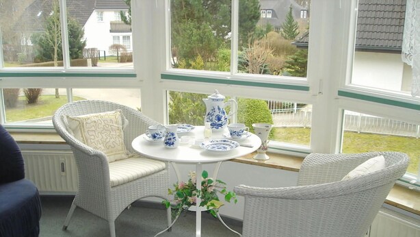 A conservatory with wicker furniture, a white table with blue and white china, and a view of a residential area. - Kurpark Residence Spot (Timmendorfer Strand)